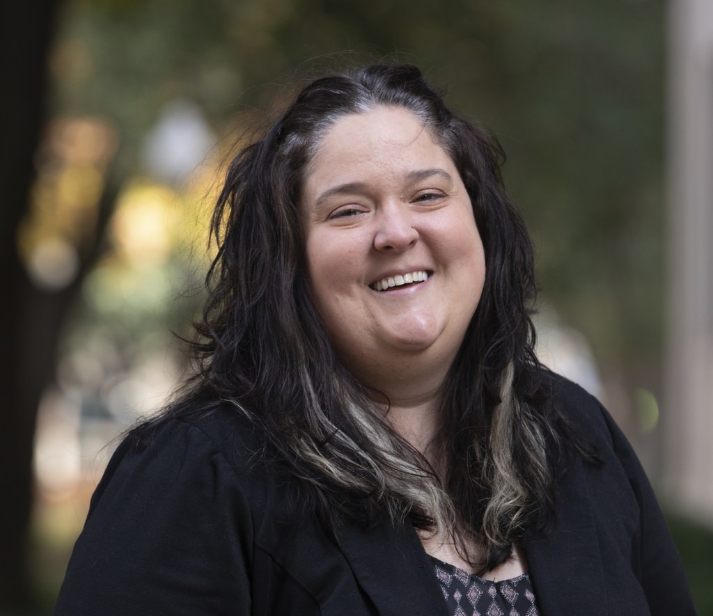Head shot of a white female with brown hair in a black suit jacket. She is smiling and in front of a group of trees. 
